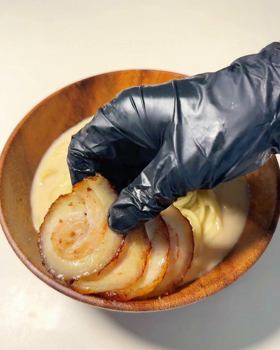 Une main gantée disposant des tranches de porc chashu grillé sur les nouilles ramen.