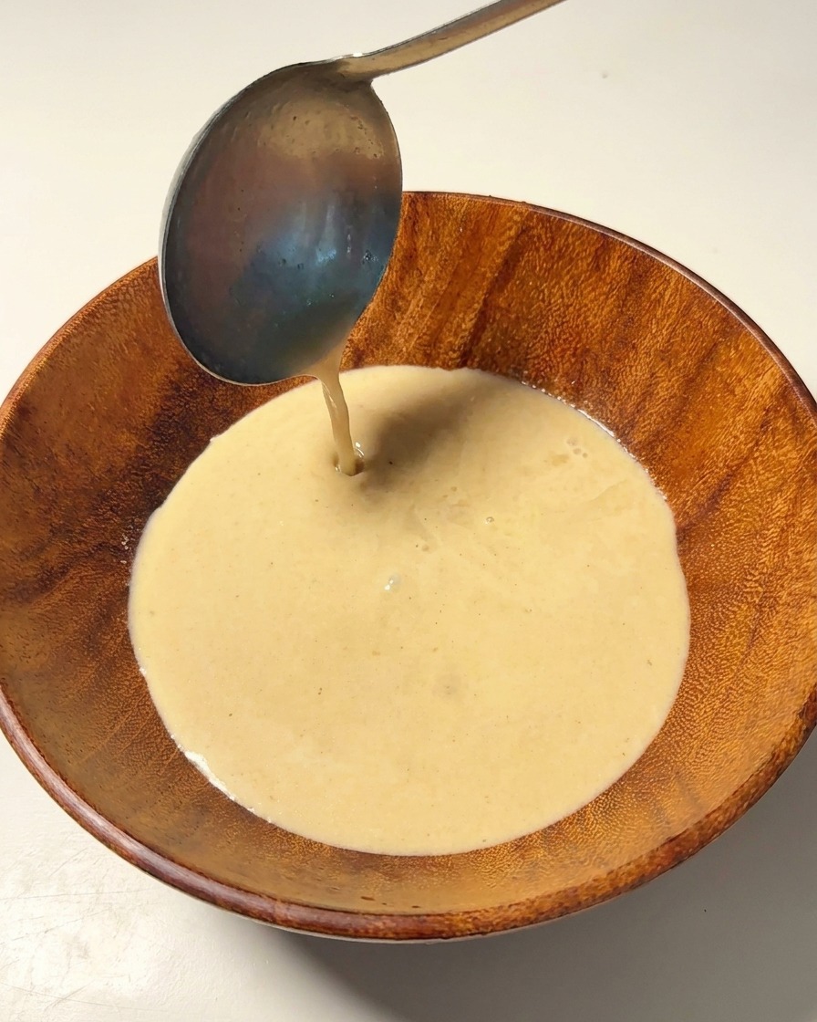 Bouillon de porc crémeux versé à laide dune louche métallique dans un bol en bois.