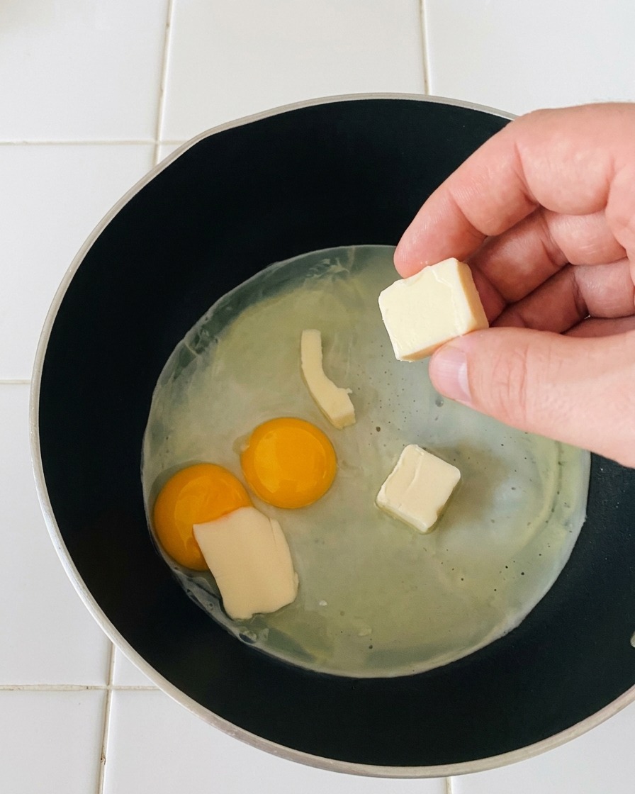 Une main déposant de petites tranches rectangulaires de mozzarella blanche sur des œufs au plat en train de cuire doucement.