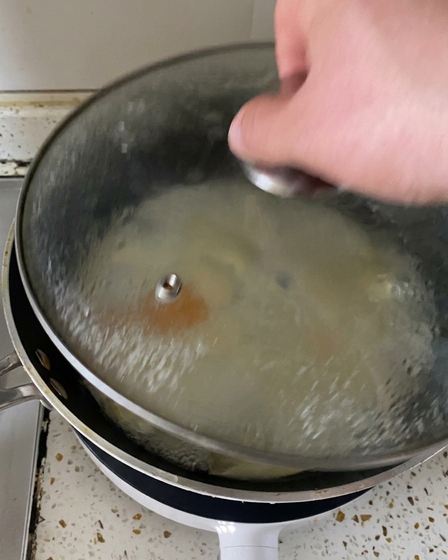 Une main soulevant un couvercle en verre dune poêle pour révéler de la vapeur et des œufs parfaitement cuits recouverts de fromage fondu.