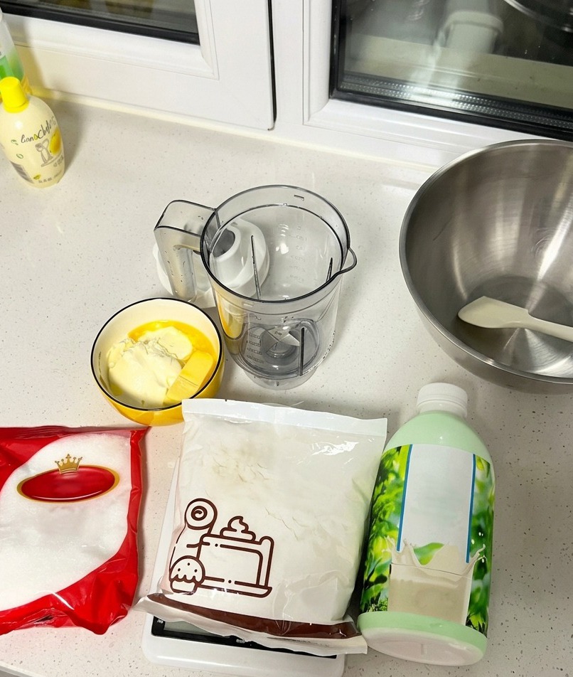 Ingrédients de pâtisserie disposés sur un plan de travail, incluant une bouteille de lait, du sucre, de la farine, du beurre et du fromage à la crème.