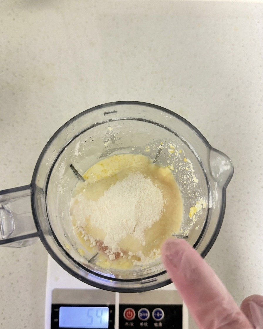 Farine et autres ingrédients secs versés sur le mélange humide dans le bol dun blender posé sur une balance de cuisine.