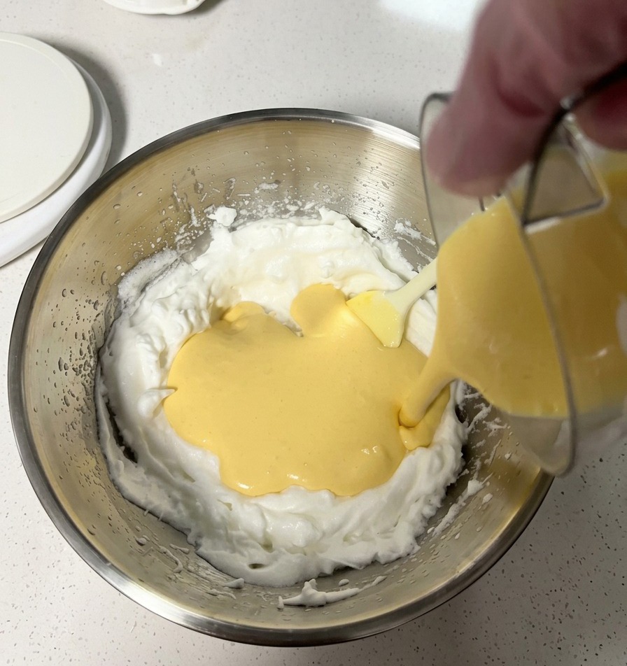 Pâte jaune et lisse versée dans un grand saladier en inox rempli de blancs dœufs en neige bien mousseux.