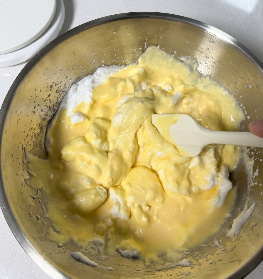 Incorporation délicate dune pâte jaune pâle avec des blancs dœufs en neige dans un saladier en inox à laide dune maryse en silicone blanche.