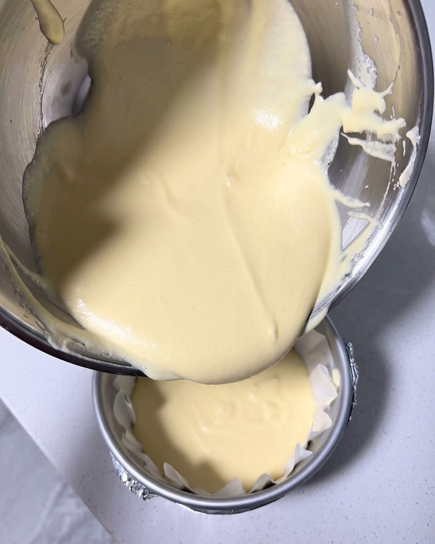 La pâte du cheesecake, lisse et jaune pâle, en train dêtre versée dans un moule rond de 15 cm chemisé de papier sulfurisé.