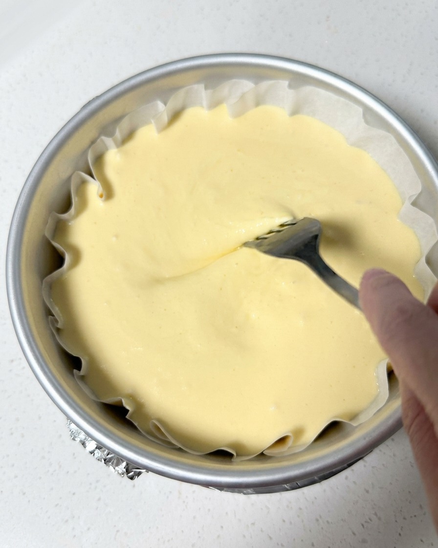 Une main utilisant une fourchette pour dessiner des tourbillons à la surface de la pâte à cheesecake dans son moule.