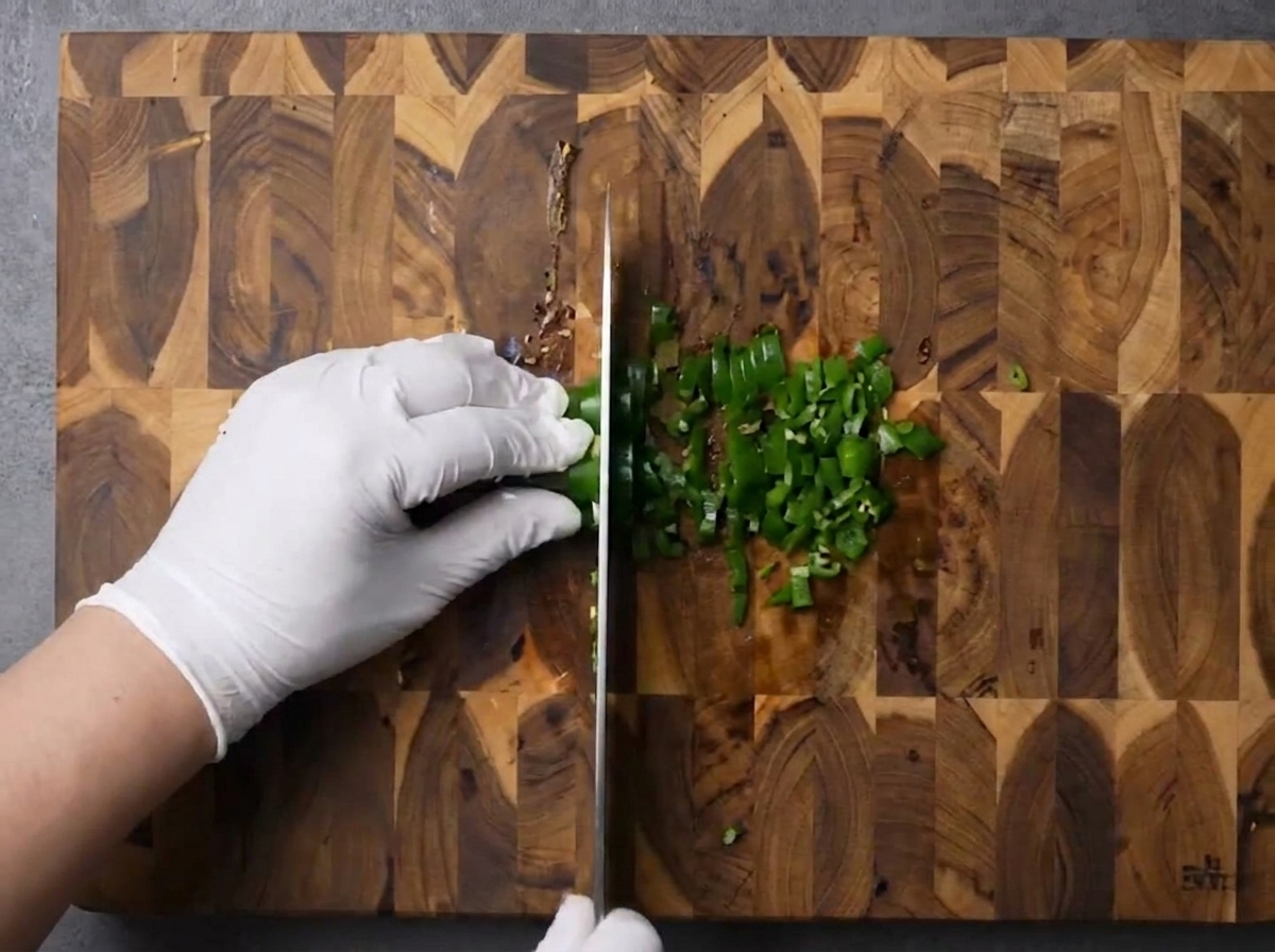 Mains gantées hachant finement des piments verts sur une planche à découper.