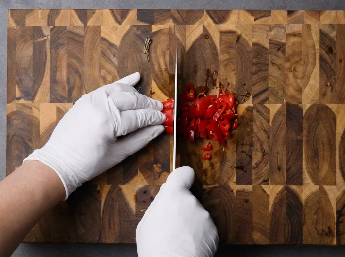 Mains gantées hachant finement des piments rouges frais sur une planche en bois.