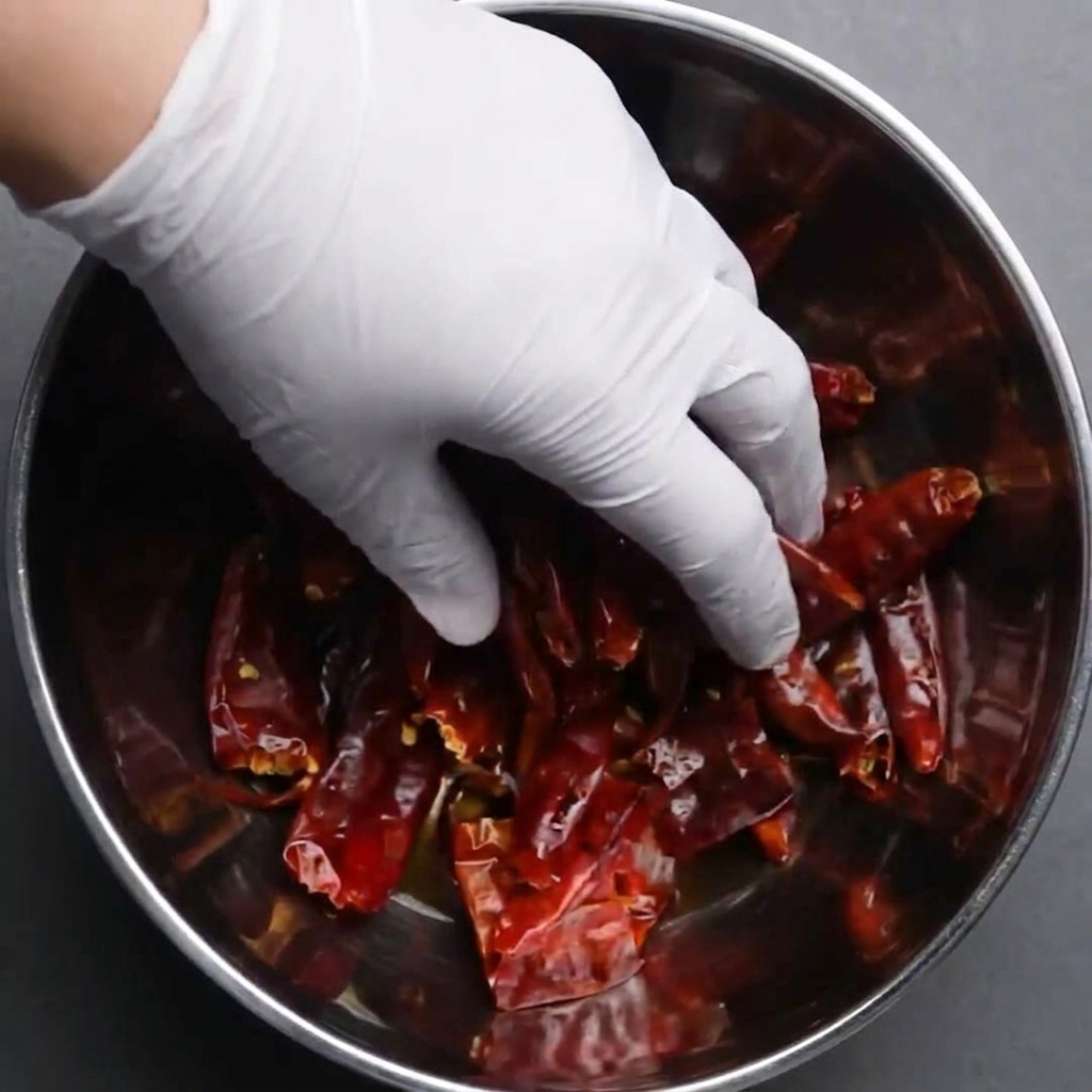 Une main gantée enfonçant des piments rouges séchés dans un bol deau pour les réhydrater.