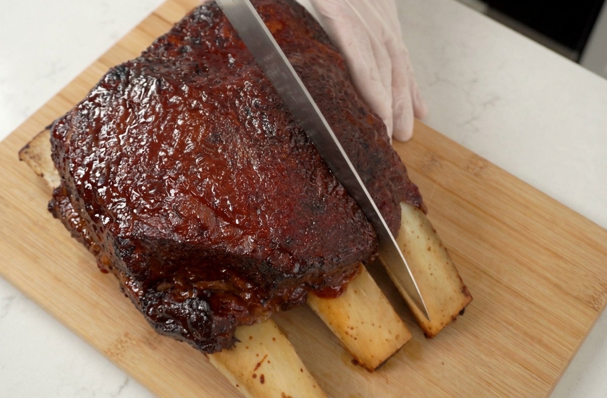 Une main gantée tenant un couteau tranchant découpant un travers de bœuf rôti et glacé.