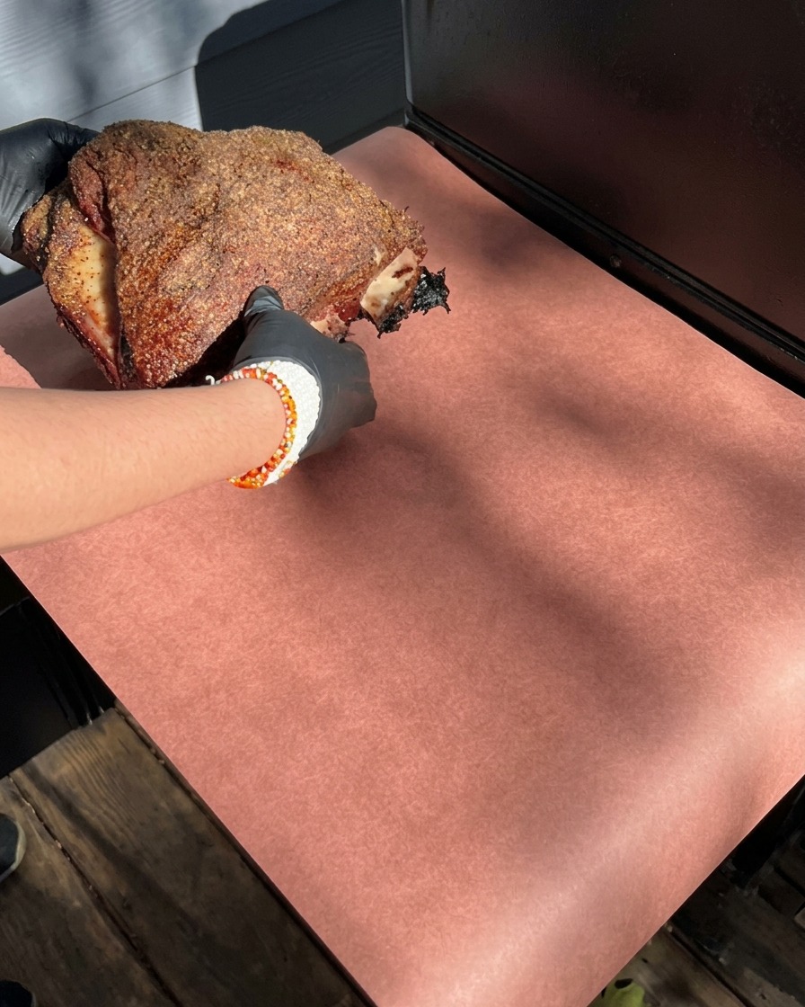 Mains gantées tenant un grand travers de bœuf fumé avec une croûte sombre, le posant sur une feuille de papier boucher rose.