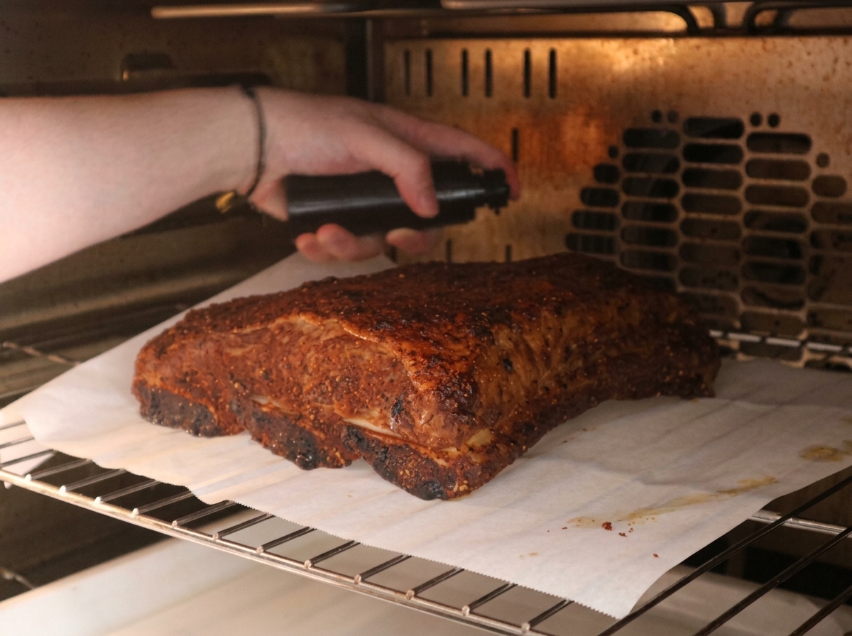 Une main tenant un vaporisateur noir, pulvérisant un mélange sur un travers de bœuf aux épices rôtissant au four.