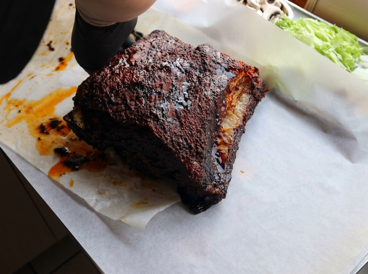 Mains portant des gants noirs plaçant un travers de bœuf rôti et sombre sur une feuille de papier sulfurisé blanc.
