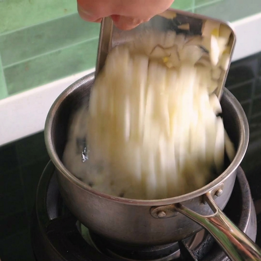 Oignons blancs coupés en dés versés depuis un racloir métallique dans une petite casserole en inox sur une cuisinière à gaz.