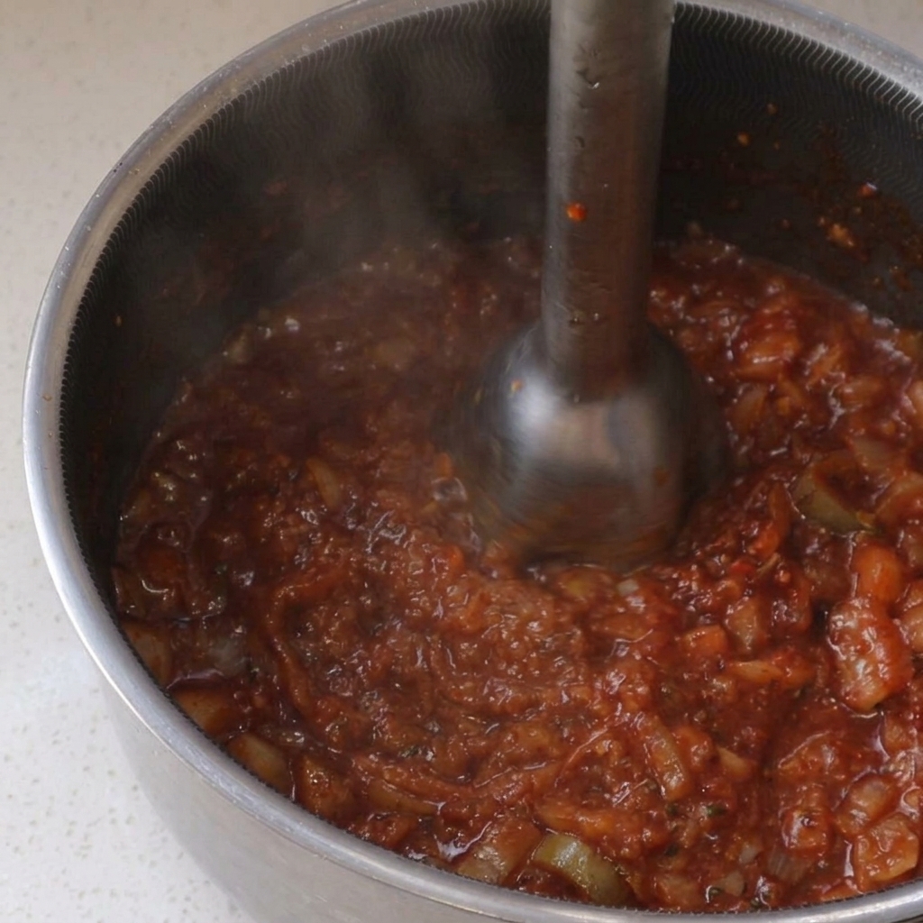 Utilisation dun mixeur plongeant métallique pour réduire en purée une sauce BBQ rouge mijotant dans une casserole en inox.
