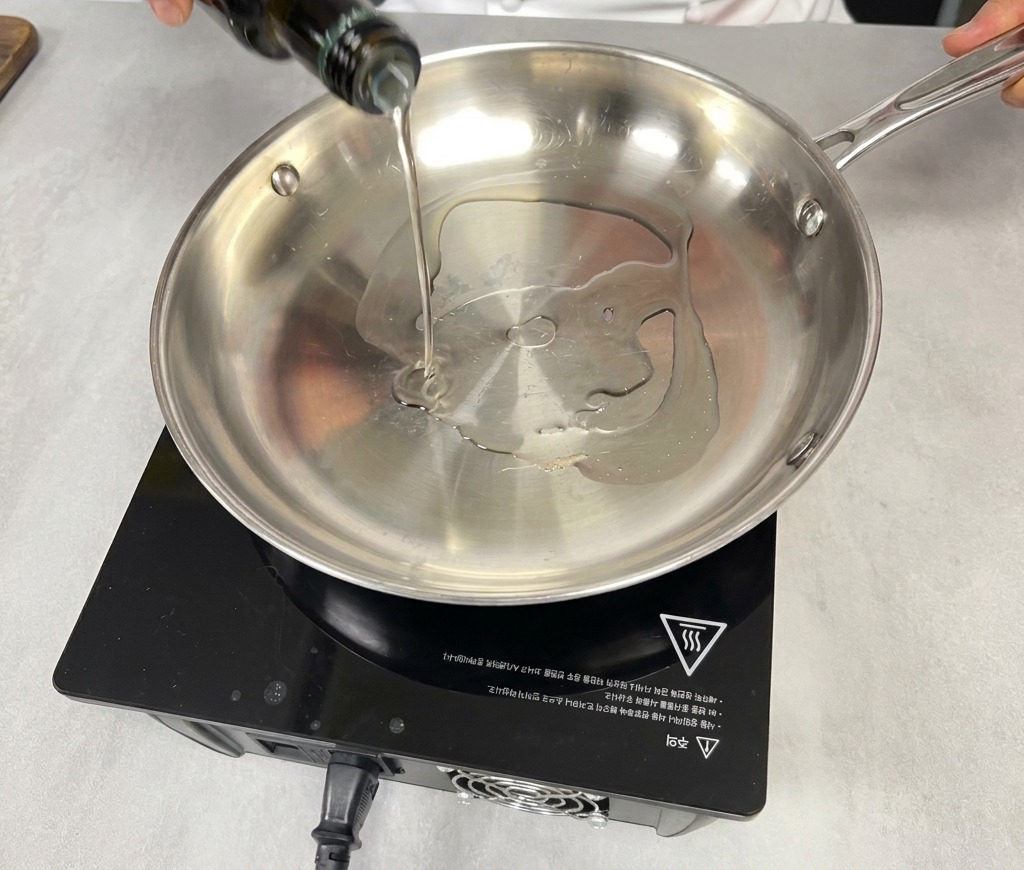 Une main versant de lhuile de cuisson dune bouteille en verre vert foncé dans une poêle en acier inoxydable vide sur une plaque à induction noire.