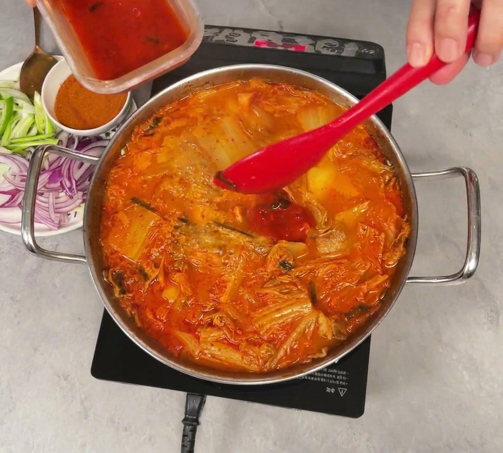 Du jus de kimchi rouge vif versé dun petit récipient en plastique dans une marmite frémissante de ragoût de kimchi et de légumes, mélangé avec une spatule rouge.