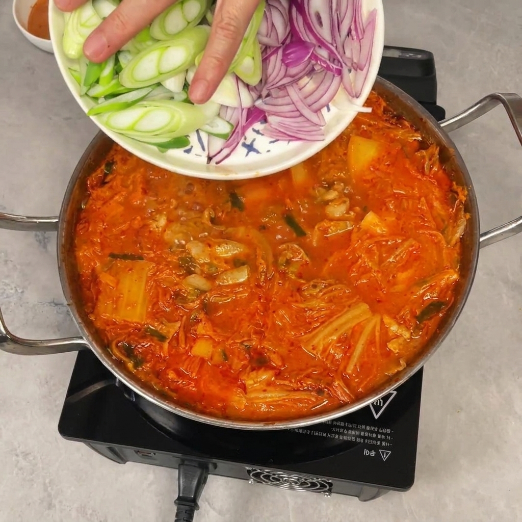 Une main faisant glisser des oignons verts et rouges émincés depuis une assiette dans une marmite frémissante de ragoût de kimchi rouge.