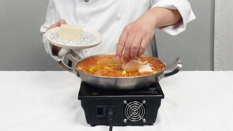 Le chef dépose délicatement des tranches carrées de tofu blanc dans une marmite frémissante de ragoût de kimchi rouge.
