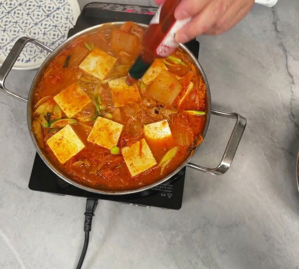 Une personne verse de la sauce pimentée Tabasco depuis une petite bouteille dans une marmite bouillonnante de ragoût de kimchi rouge garnie de cubes de tofu blanc.
