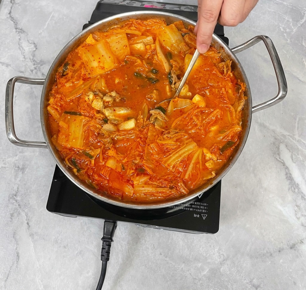 Une main utilisant une cuillère en acier inoxydable pour remuer délicatement un ragoût de kimchi rouge vif, mélangeant les assaisonnements au bouillon.