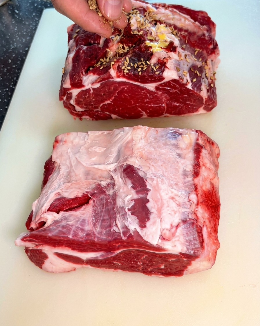 Une main saupoudrant des graines de fenouil sur deux larges morceaux de paleron de bœuf cru.