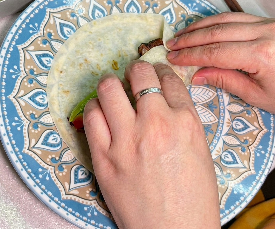 Deux mains roulant soigneusement une tortilla garnie de bœuf et de légumes.