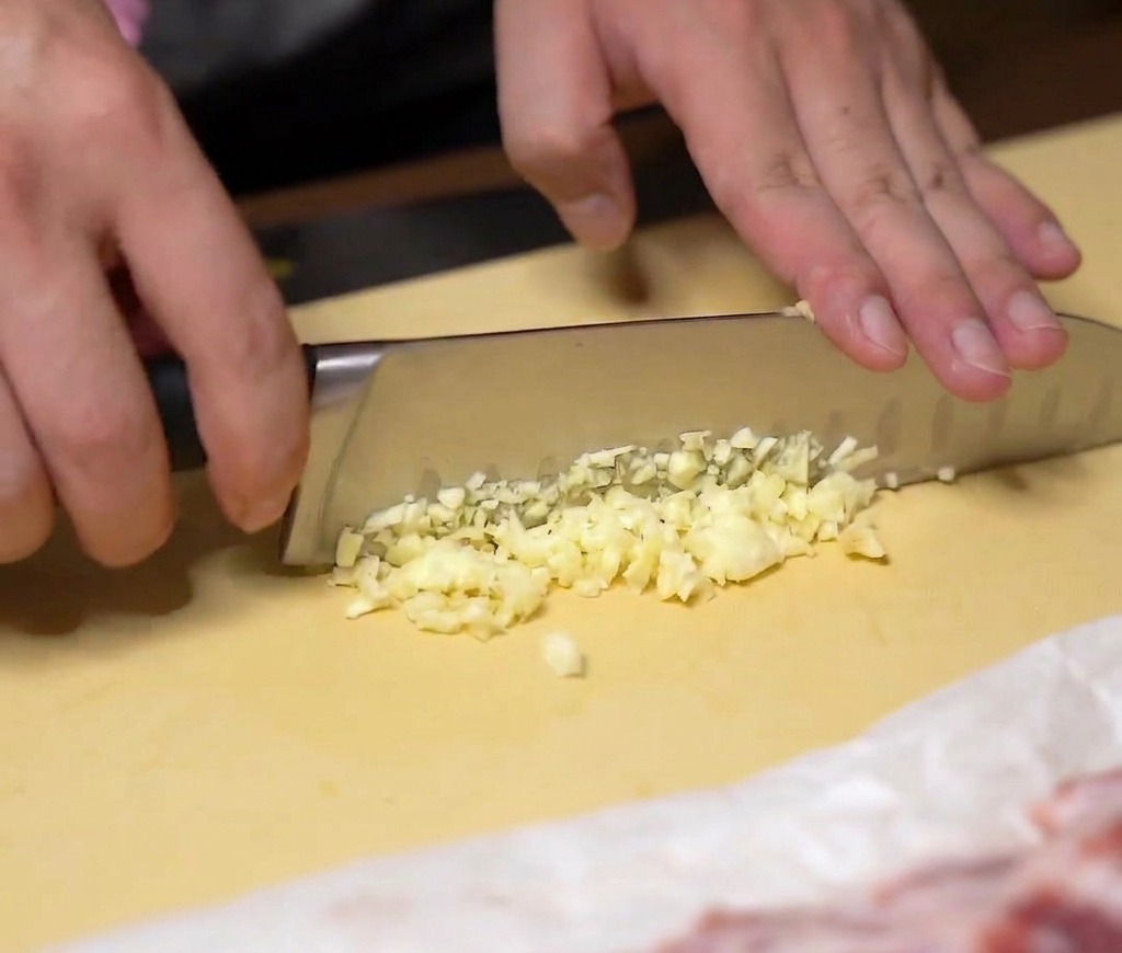Mains hachant finement des gousses dail frais avec un couteau de chef sur une planche jaune.