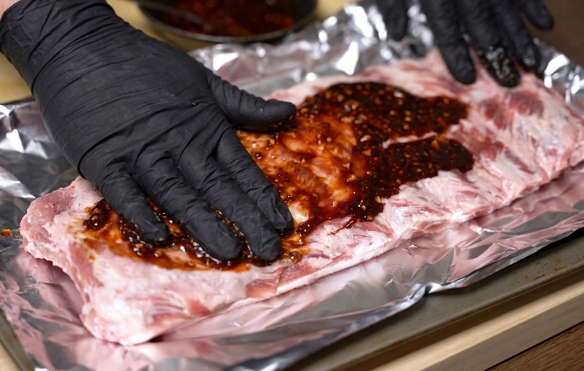 Mains gantées frottant une marinade rouge épaisse sur un travers de porc cru posé sur du papier aluminium.