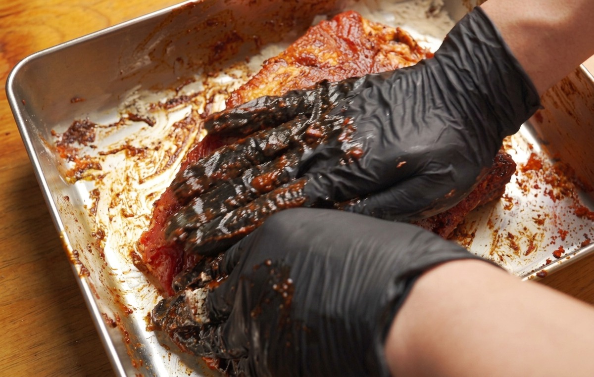 Mains gantées frottant fermement une pâte de marinade brun-rouge sur un grand morceau de porc.