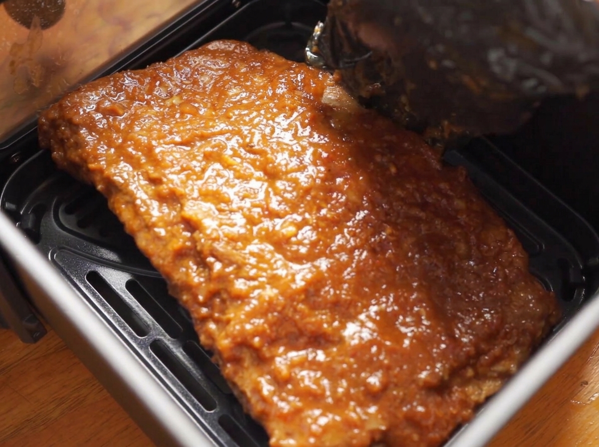 Main gantée plaçant une grande pièce de porc nappée de sauce brillante dans le panier dun air fryer.