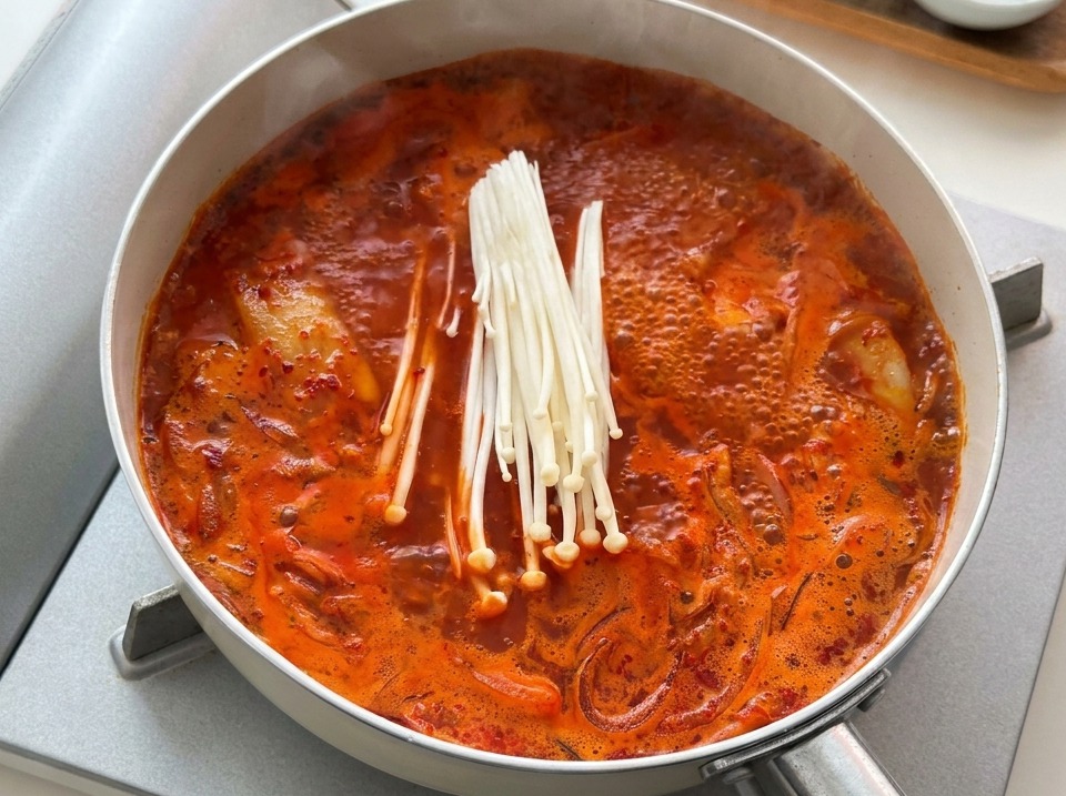 Une botte de champignons enoki blancs crus ajoutée à une casserole de ragoût de kimchi rouge bouillant.