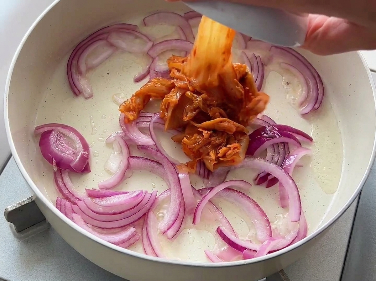 Du kimchi haché ajouté depuis un petit bol dans une poêle contenant des oignons rouges sautés.