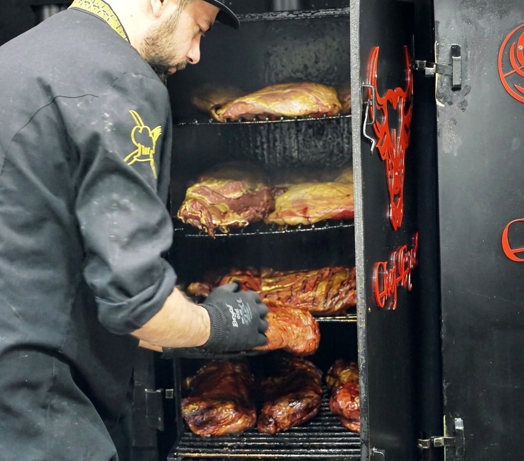 Un chef retirant une grande pièce de bœuf fumé avec une croûte sombre dun fumoir à plusieurs étages.