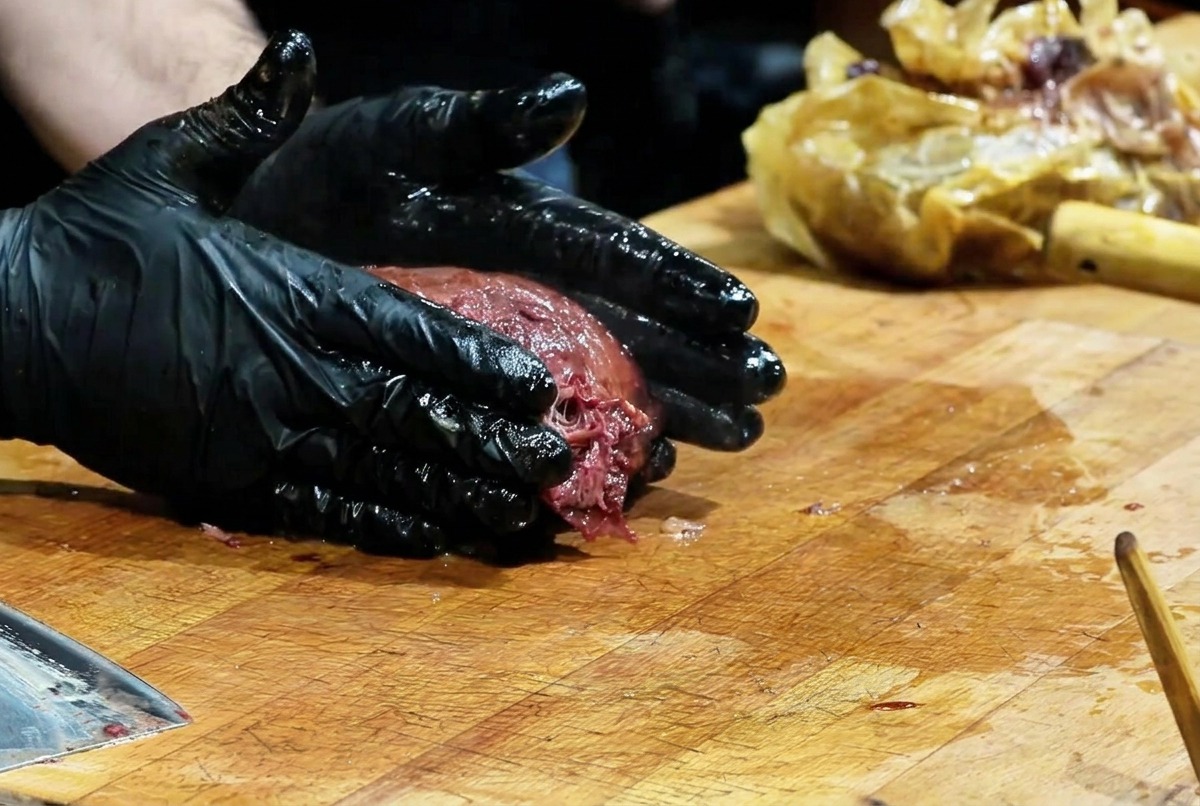 Un chef portant des gants noirs pressant une grande brisket fumée sur une planche en bois pour effilocher la viande