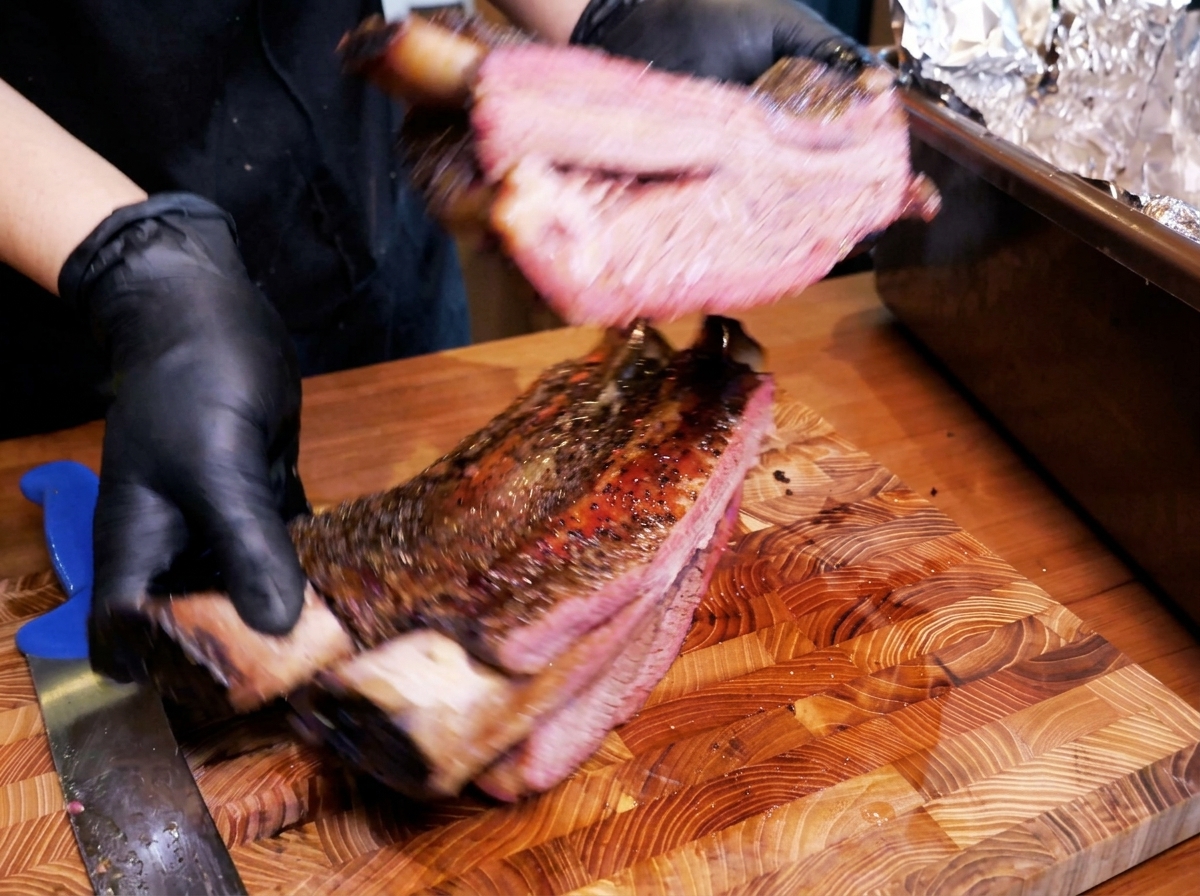 Un grand rack de côtes de bœuf fumées avec une croûte sombre épaisse, manipulé par des mains gantées, révélant un intérieur rose juteux.