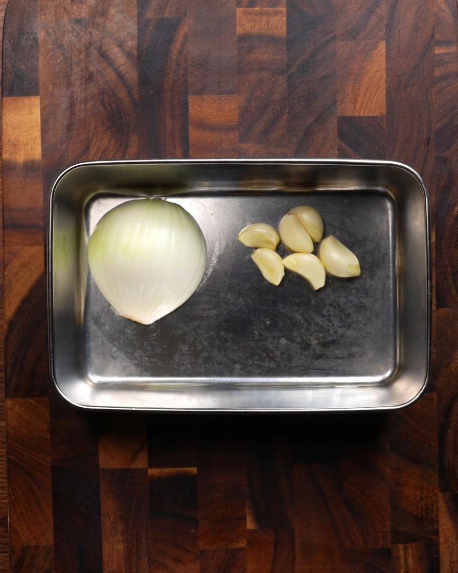 Un plateau en acier inoxydable contenant un demi-oignon blanc et plusieurs gousses dail épluchées sur une planche en bois.