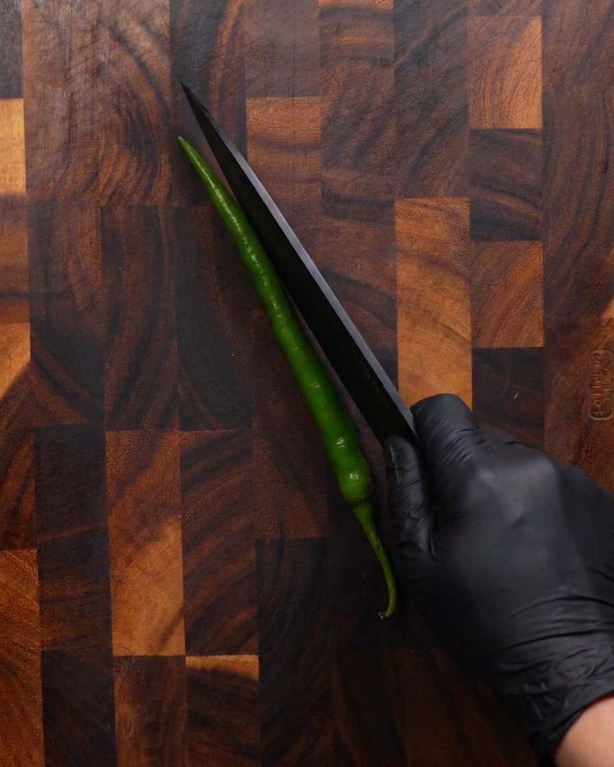 Une main gantée de noir utilise un couteau noir tranchant pour couper un long piment vert en diagonale sur une planche à découper en bois.