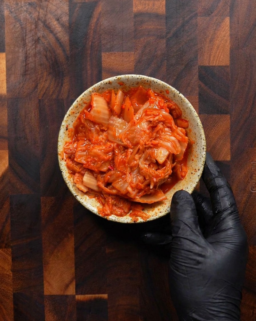 Une main gantée tenant un petit bol rustique moucheté rempli à ras bord de kimchi fermenté haché dun rouge vif.