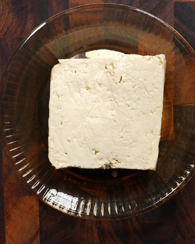 Un bloc carré de tofu blanc ferme reposant dans un bol en verre peu profond sur une surface en bois.