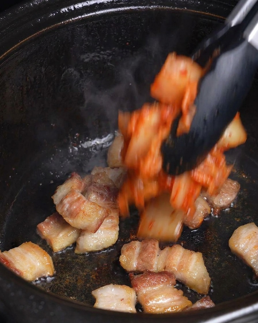 Du kimchi rouge haché est ajouté à la poitrine de porc dorée dans une marmite noire à laide de pinces.