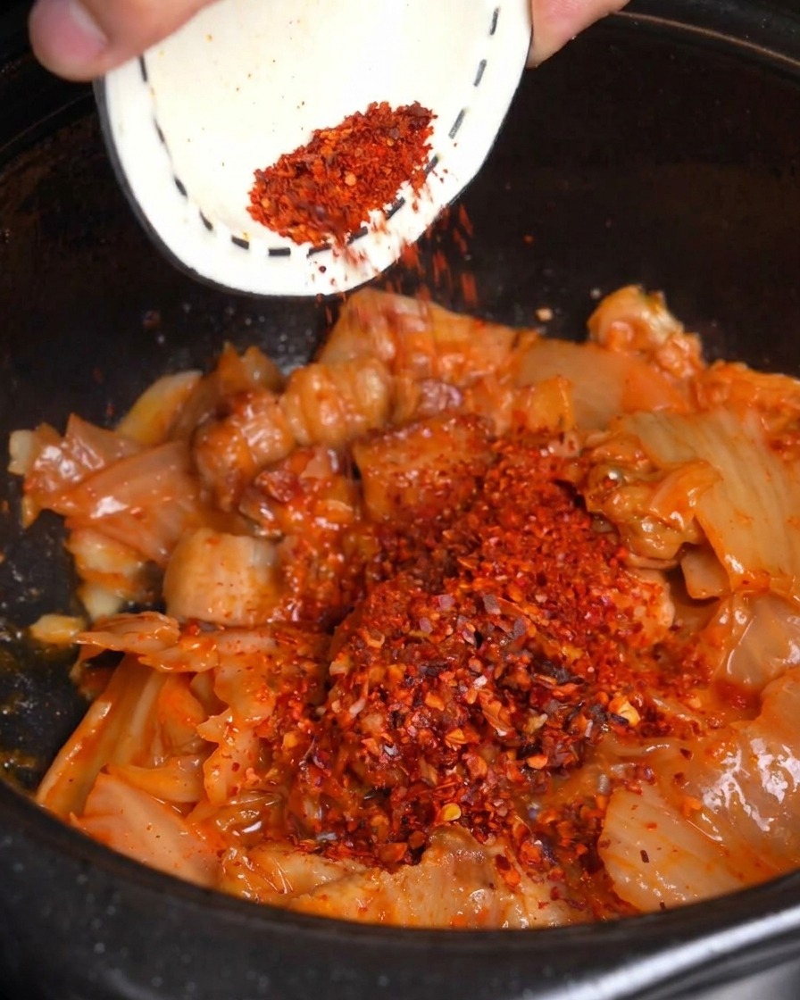 De la poudre de piment rouge est versée depuis un petit bol sur le kimchi et la poitrine de porc sautés dans une marmite noire.