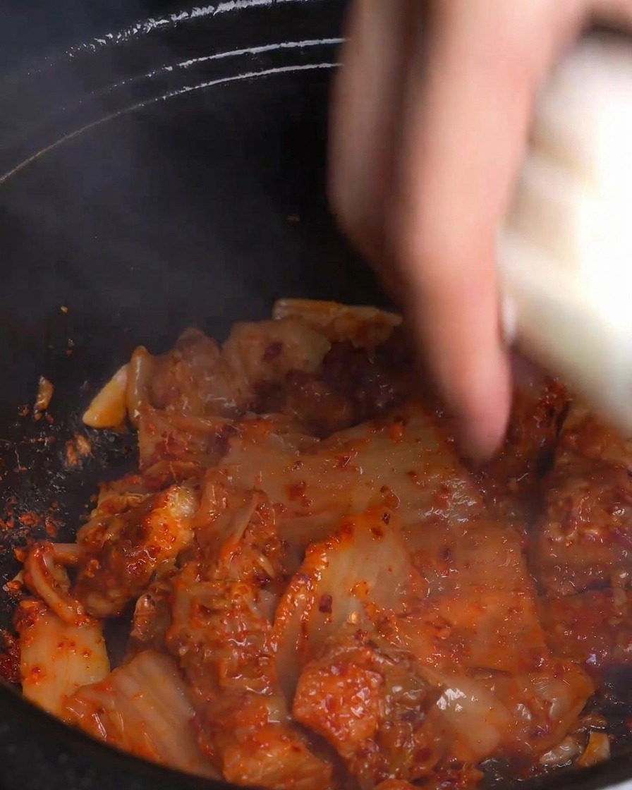 Une main jetant dépaisses rondelles doignon blanc frais dans une marmite fumante de kimchi et de poitrine de porc sautés.