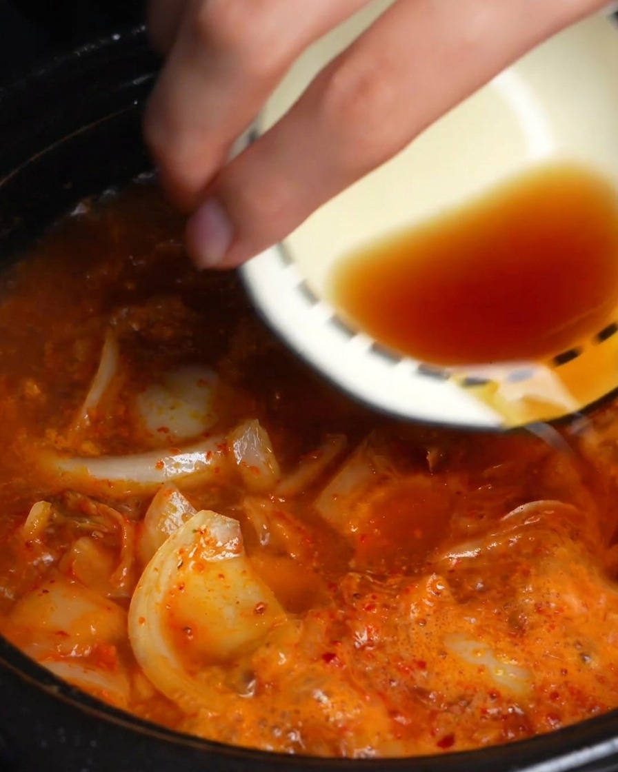 Une main versant de la sauce soja foncée et de la sauce de poisson depuis un petit bol dans un bouillon de kimchi rouge frémissant.