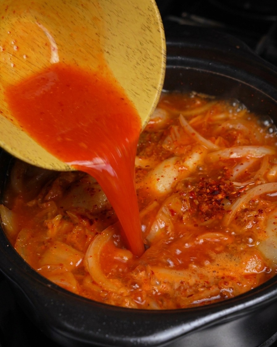 Verser le jus de kimchi rouge vif dun bol en bois dans une marmite de ragoût de porc et de kimchi bouillonnant.
