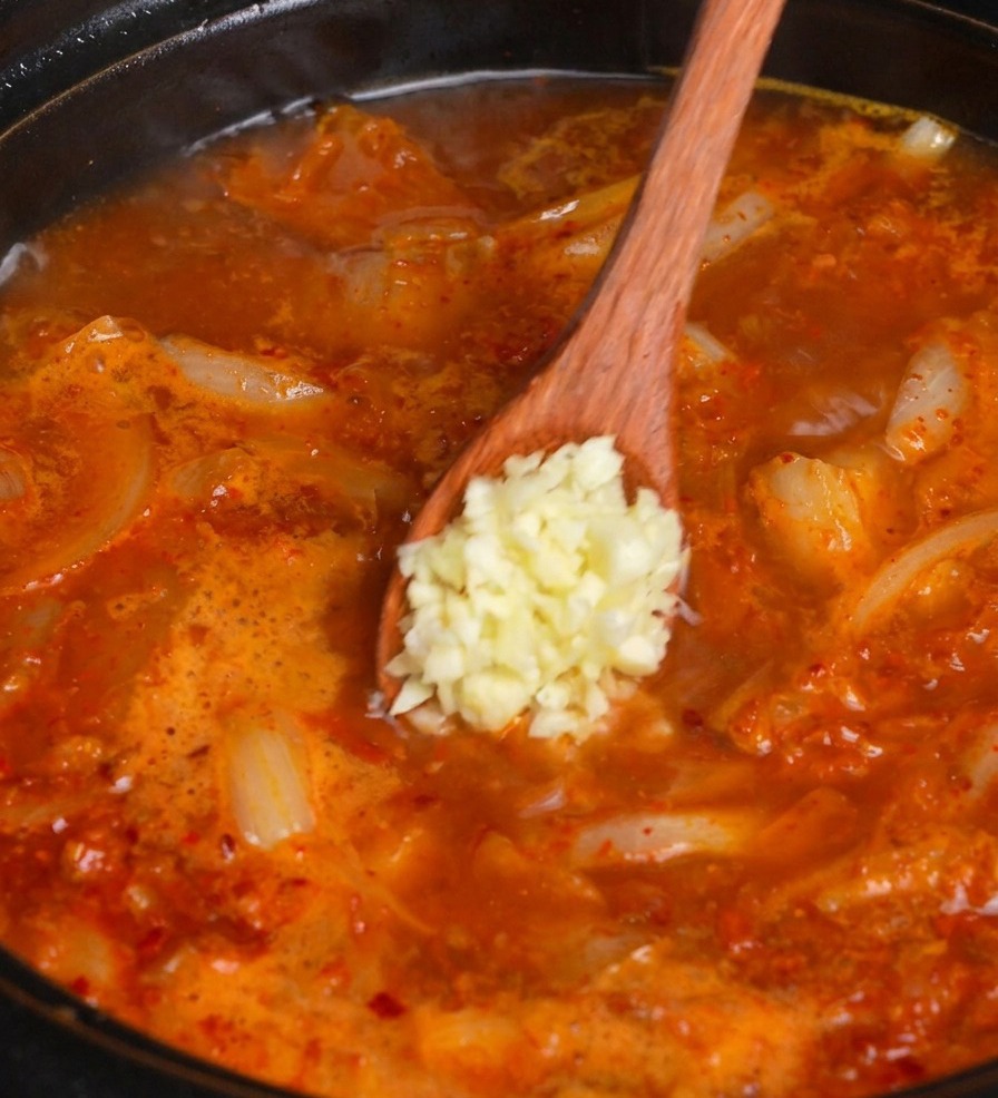 Une cuillère en bois ajoutant une cuillerée dail frais haché dans une marmite frémissante de soupe rouge au kimchi.