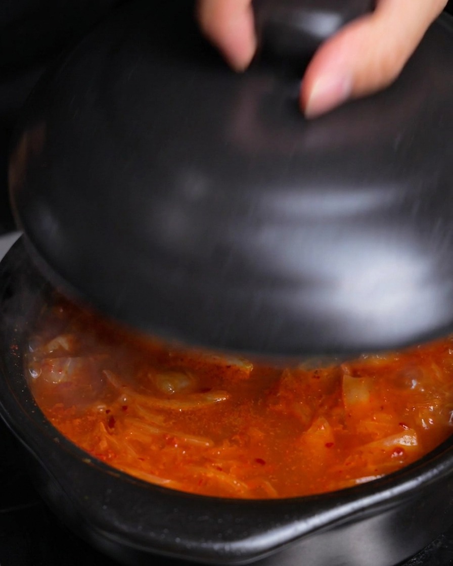 Une main plaçant un couvercle en céramique noire sur une marmite de ragoût de kimchi chaud et bouillonnant pour le laisser mijoter.