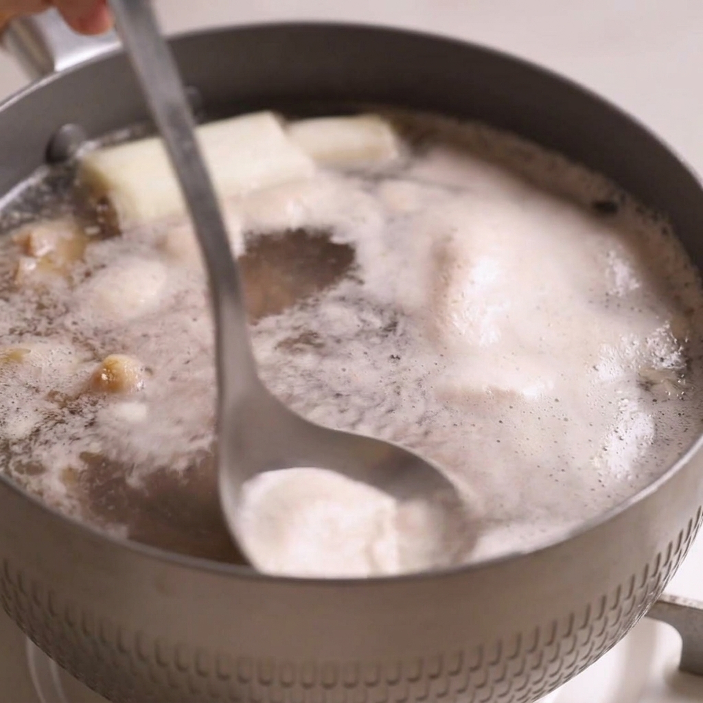 Une louche en métal plongeant dans un bouillon de bœuf bouillant pour retirer lécume grise à la surface.