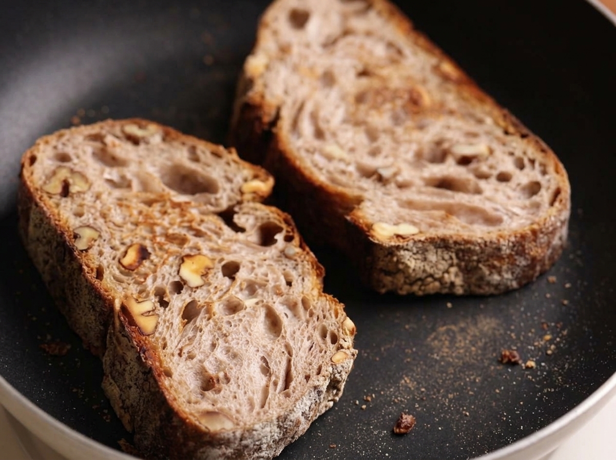 Deux tranches de pain artisanal rustique grillant dans une poêle plate sombre.