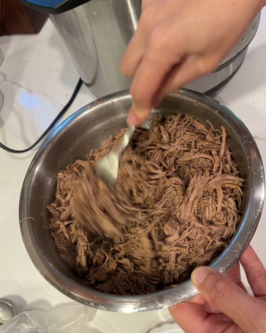 Deux mains utilisant des fourchettes pour effilocher le bœuf tendre dans un grand bol inox.